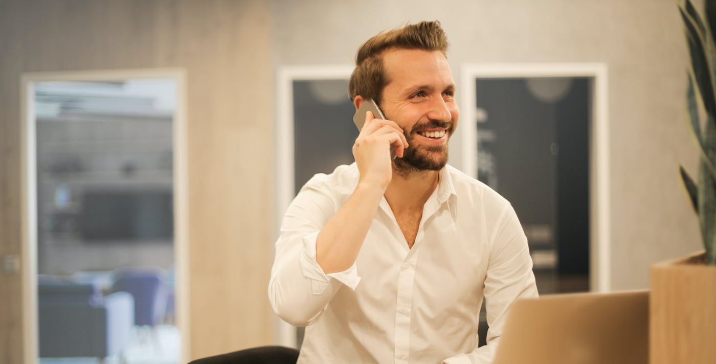 Man in white shirt sits at work bench with his laptop and speaks on mobile phone